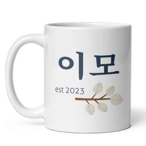 May include: White ceramic mug with the Korean text "이모" and the text "est 2023" below. A brown twig with beige leaves is on the right side of the mug.