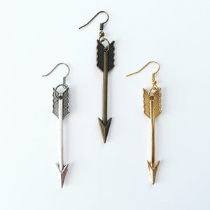 May include: Three arrow-shaped earrings in silver, bronze, and gold tones. Each earring features a hook for pierced ears and a detailed arrow design. The earrings are displayed against a white background.