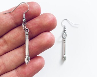 May include: Silver-colored earrings in the shape of paintbrushes. Each earring features a hook, a cylindrical handle with etched details, and a brush tip. The earrings are displayed against a white background.