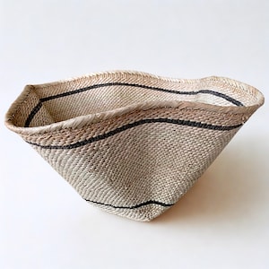 May include: A handwoven basket with a unique, asymmetrical shape. The basket is made of natural fibers in a light beige color, with dark gray stripes. The rim is reinforced with a woven border.