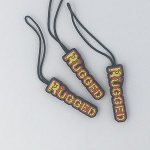 以下が含まれることがあります： 「RUGGED」の文字が黄色と赤で書かれた、黒いジッパープルが3つ。各プルには、取り付け用の黒いコードループが付いています。プルは白い背景にあります。