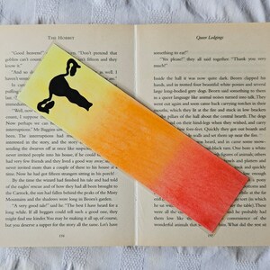 May include: A watercolor bookmark with a black cat silhouette on a yellow, orange, and red gradient background. The bookmark is placed on a page of a book titled "The Hobbit".
