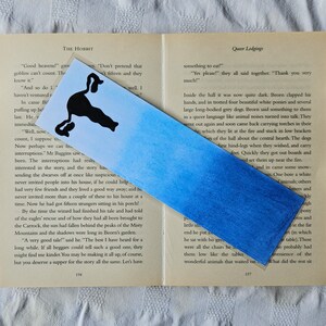 Puede incluir: Un marcap&aacute;ginas de acuarela azul y negro con una silueta de un gato. El marcap&aacute;ginas tiene un degradado de tonos azules, comenzando con un azul oscuro en la parte inferior y desvaneci&eacute;ndose a un azul m&aacute;s claro en la parte superior. La silueta del gato es negra y est&aacute; colocada en el medio del marcap&aacute;ginas. El marcap&aacute;ginas es una excelente manera de marcar tu lugar en un libro.