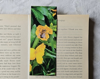 May include: A rectangular bookmark featuring a vibrant image of a bee on a yellow flower, set against a backdrop of green foliage. The bookmark is placed within the pages of a book.