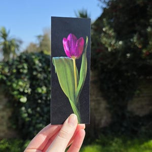 May include: A bookmark depicting a purple tulip with a yellow stem and green leaves against a black backdrop. The bookmark is held in a hand, with a blurred outdoor scene visible in the background.