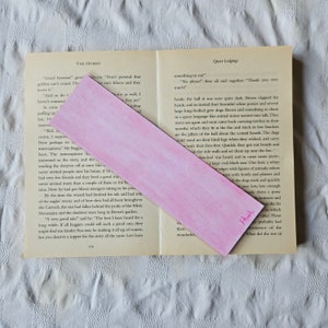 May include: A pink bookmark with a watercolor design. The bookmark is folded in half and has the word "Thanks" written on it in pink ink.