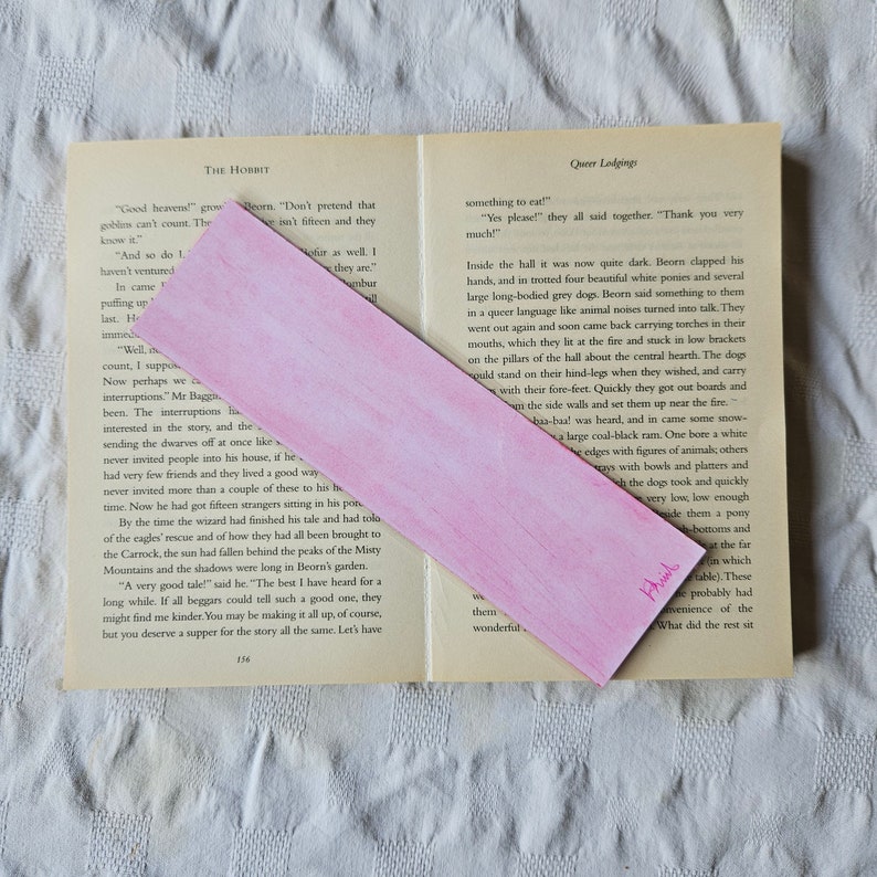 May include: A pink bookmark with a watercolor design. The bookmark is folded in half and has the word "Thanks" written on it in pink ink.