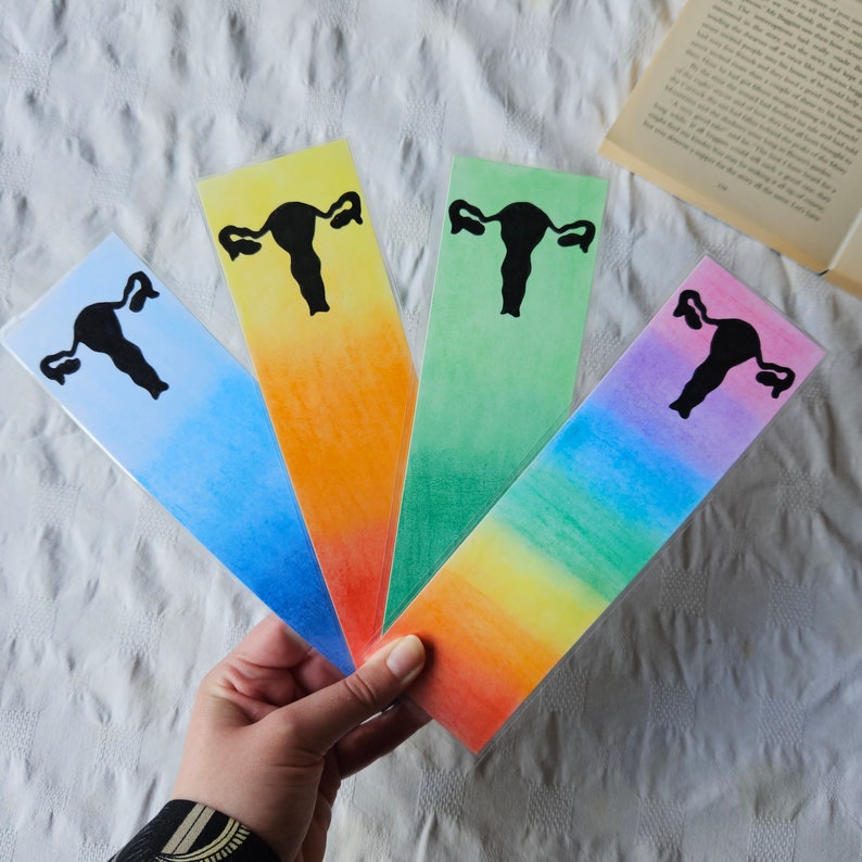 May include: Four colorful bookmarks with a black silhouette of a uterus on each. The bookmarks are in shades of blue, orange, green, and pink.