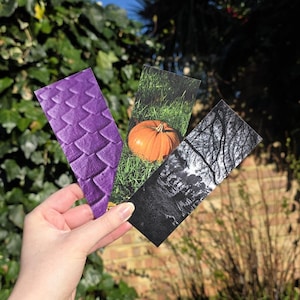 May include: Three rectangular bookmarks are held up against a green leafy background. One bookmark has a purple scale pattern, another features an orange pumpkin in grass, and the third is a black and white image of trees.