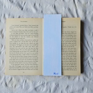 Puede incluir: Un primer plano de un libro abierto con un marcap&aacute;ginas azul. El libro se titula "El Hobbit" y el texto de la p&aacute;gina describe una escena con enanos y goblins. El marcap&aacute;ginas tiene la palabra "Phius" escrita en &eacute;l.
