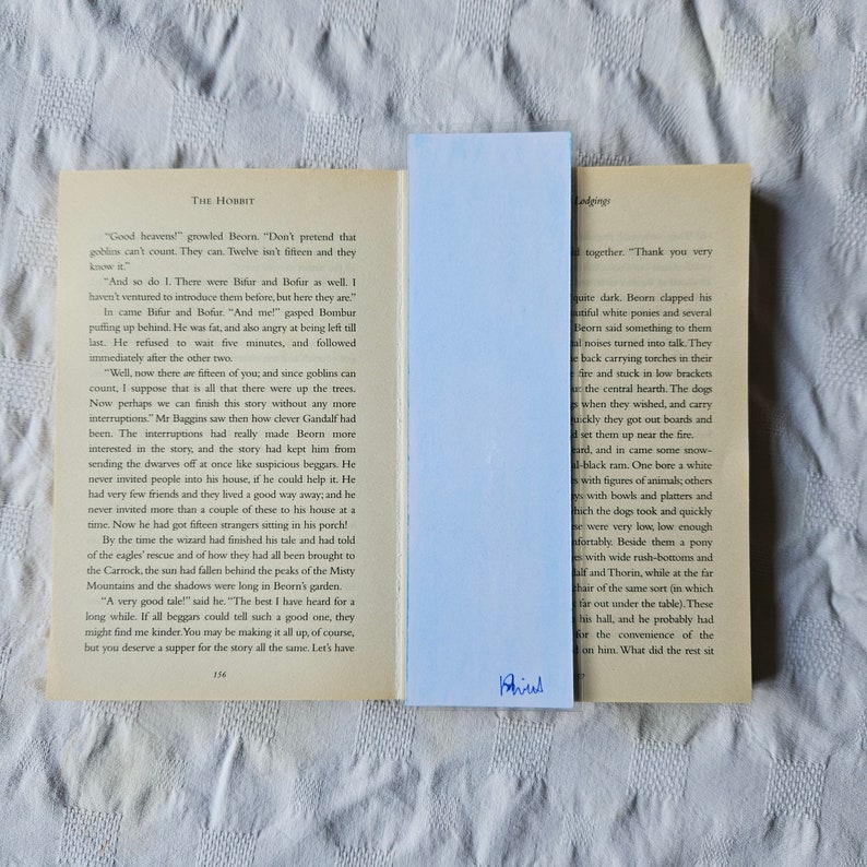 Puede incluir: Un primer plano de un libro abierto con un marcap&aacute;ginas azul. El libro se titula "El Hobbit" y el texto de la p&aacute;gina describe una escena con enanos y goblins. El marcap&aacute;ginas tiene la palabra "Phius" escrita en &eacute;l.