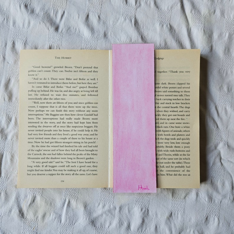 May include: A close-up of a book titled "The Hobbit" with a pink bookmark. The bookmark has the word "Khinh" written on it. The book is open to a page with text about a group of dwarves and a wizard named Gandalf.