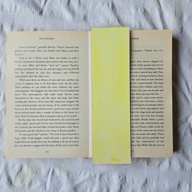 May include: A close-up of a book page with text from the novel "The Hobbit" by J.R.R. Tolkien. The text describes a scene where the character Beorn is hosting a group of dwarves for dinner. The page is yellowed with age and has a worn, vintage look.