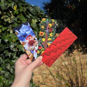 Könnte beinhalten: Drei farbenfrohe Lesezeichen werden vor dem Hintergrund von grünem Laub und blauem Himmel präsentiert. Eines zeigt Gebäck mit Marmelade, ein anderes ein Schwert in Blumen und das dritte ein rotes Schuppenmuster.