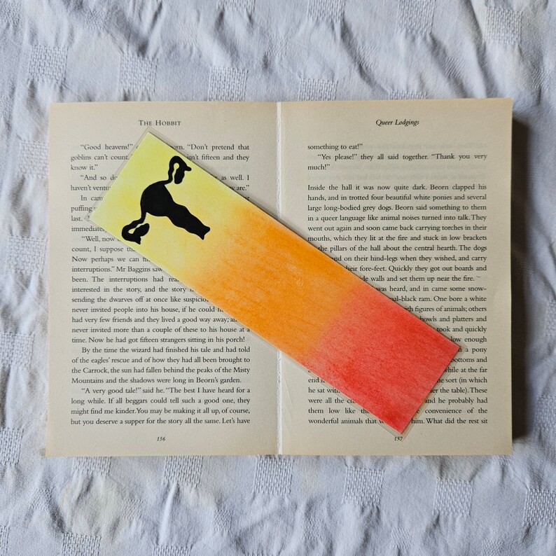 May include: A watercolor bookmark with a black cat silhouette on a yellow, orange, and red gradient background. The bookmark is placed on a page of a book titled "The Hobbit".