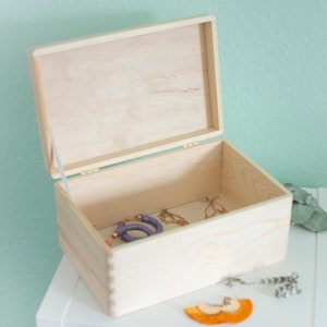 Könnte beinhalten: Eine Holzkiste mit einem Scharnierdeckel, geöffnet, um ein violettes Perlenarmband und goldenen Schmuck im Inneren zu zeigen. Die Kiste ist aus hellem Holz und hat eine natürliche Oberfläche.