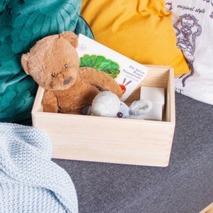 Könnte beinhalten: Eine Holzkiste mit einem braunen Teddybär, einem grauen Stofftier und einem Kinderbuch mit dem Titel "Die kleine Raupe Nimmersatt" von Eric Carle.