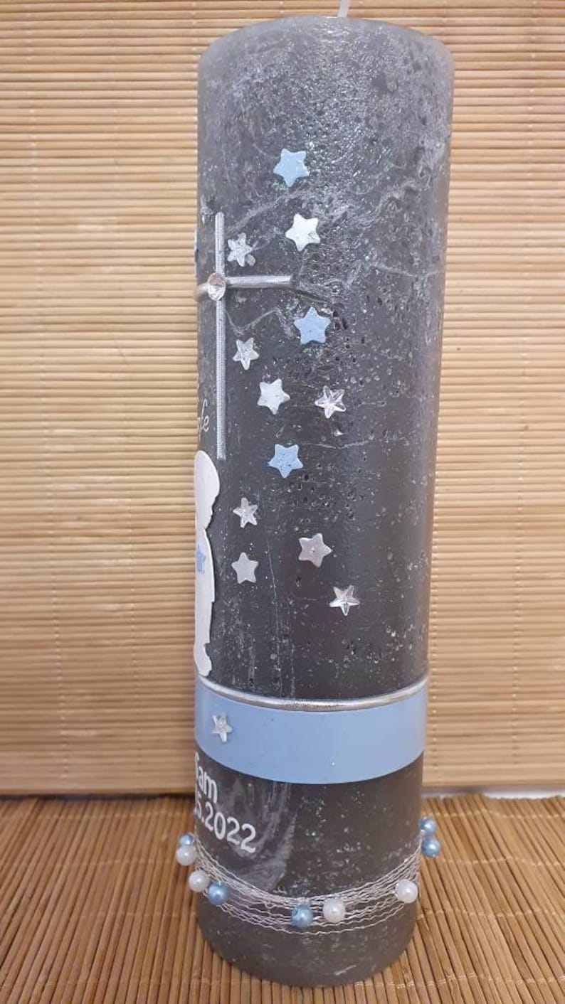 Pu&ograve; includere: Una candela grigia con un nastro blu e argento. La candela &egrave; decorata con stelle argentate e una silhouette bianca di un bambino. Il testo "Sam 5.2022" &egrave; stampato sulla candela.