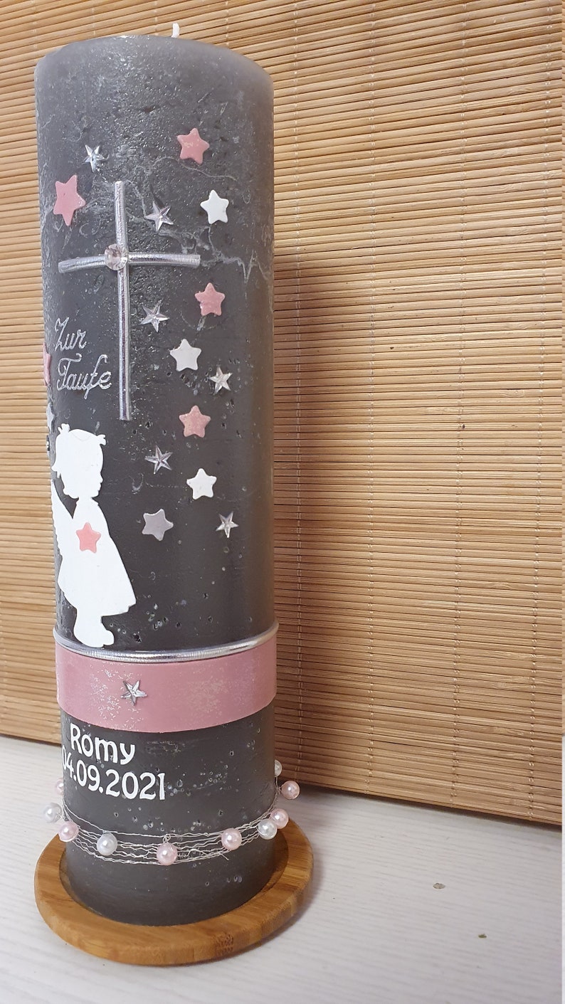 Pu&ograve; includere: Una candela battesimale grigia con un nastro rosa e un bordo di perle bianche. La candela presenta una silhouette bianca di un angelo con stelle rosa e una croce. Il testo "Zur Taufe" e "Romy 04.09.2021" &egrave; scritto sulla candela.