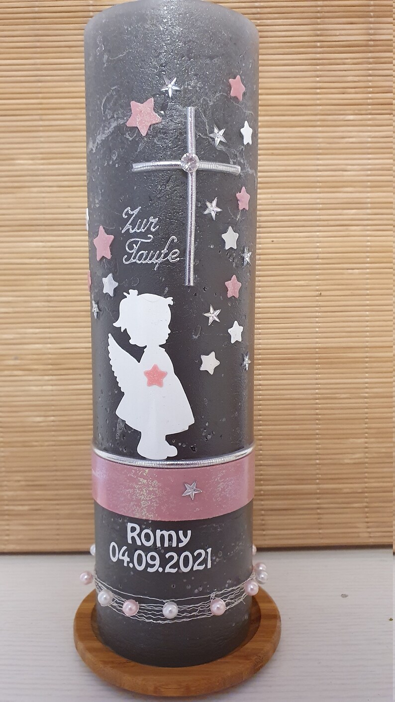 Pu&ograve; includere: Una candela grigia con una silhouette di angelo rosa e bianca, una croce e stelle. La candela ha un nastro rosa con il testo "Zur Taufe" e "Romy 04.09.2021".