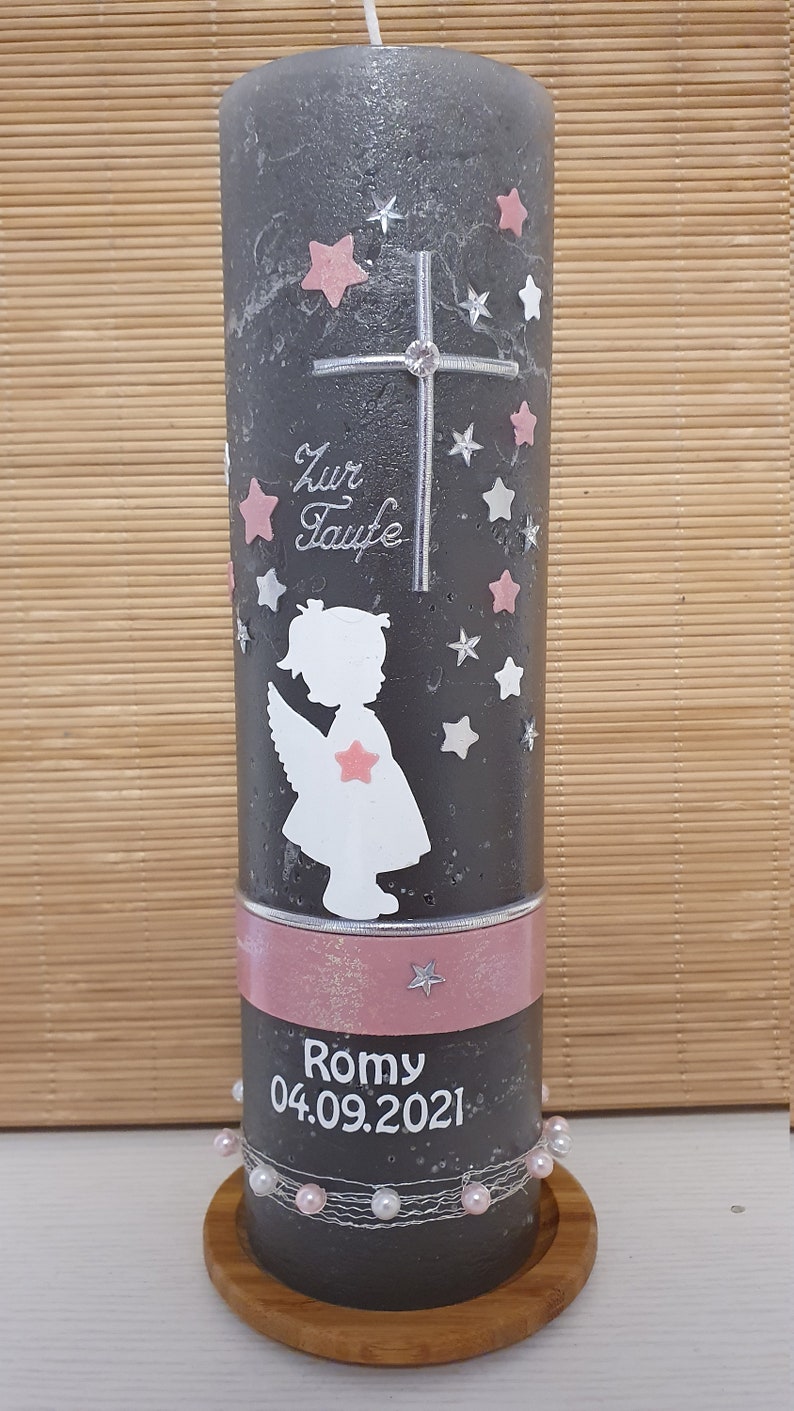 Pu&ograve; includere: Una candela battesimale grigia con un motivo a stelle rosa e bianche. La candela ha una croce argentata con un strass, una silhouette bianca di un angelo con ali rosa e il testo "Zur Taufe" in lettere argentate. La candela ha anche un nastro rosa con il nome "Romy" e la data "04.09.2021" in lettere bianche.