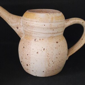May include: A ceramic creamer or small pitcher with a rounded body, a narrow neck, and a wide spout. The vessel features a speckled cream and tan glaze with horizontal bands around the neck and a curved handle extending from the side of the body.