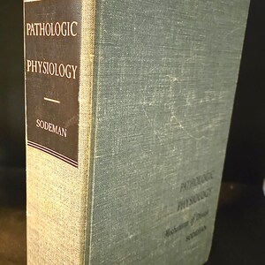 May include: A hardcover book titled Pathologic Physiology by Sodeman with a textured light blue-grey cloth cover. The spine features a black rectangular panel with white text reading Pathologic Physiology and Sodeman, with gold lettering at the base.