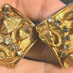 Puede incluir: Un par de pendientes cuadrados de metal dorado con una superficie texturizada, motivos de hojas en relieve y una línea curva de pequeños diamantes de imitación transparentes incrustados en el centro de cada pieza, creando un diseño botánico.
