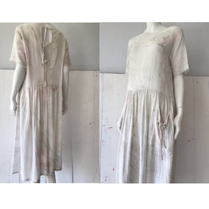 May include: A white linen dress with a tie at the back and a loose, flowing silhouette. The dress has a distressed look with paint splatters and a pocket on the side.