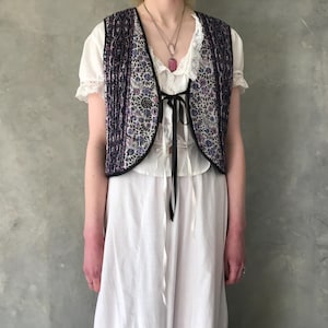 Puede incluir: Una blusa blanca sin mangas con un chaleco estampado floral negro. El chaleco tiene un lazo en la parte delantera y un ribete de encaje negro.