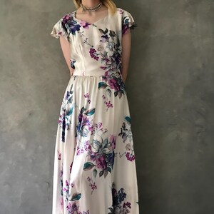 May include: A white maxi dress with a floral print featuring purple, blue, and green flowers. The dress has a scoop neck and short sleeves.