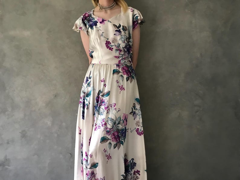 May include: A white maxi dress with a floral print featuring purple, blue, and green flowers. The dress has a scoop neck and short sleeves.