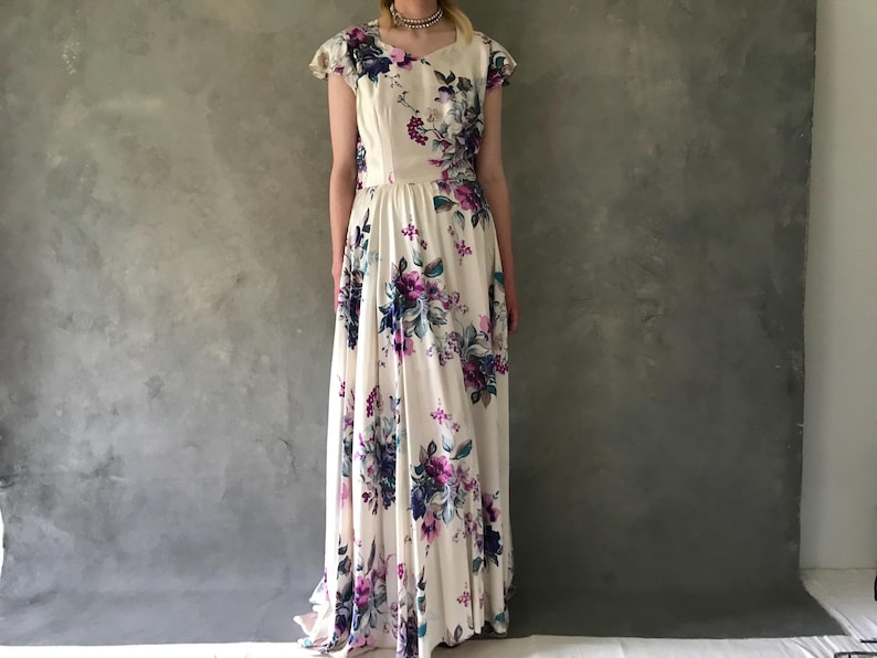 May include: A white maxi dress with a floral print in shades of purple, blue, and pink. The dress has a scoop neckline, short sleeves, and a fitted bodice. The skirt is full and flowing.