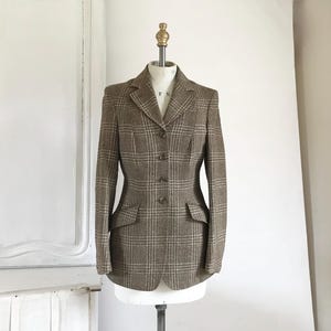 May include: A brown and beige checked blazer with a notched lapel, two front pockets, and a four-button fastening. The jacket is displayed on a mannequin against a white backdrop. The blazer has a classic, structured design.