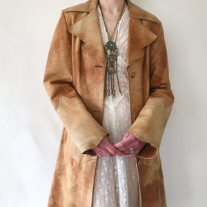 May include: A tan suede coat with a long, tailored silhouette. The coat features a notched collar, button closures, and decorative stitching. The coat is worn over a white lace dress and a long necklace.