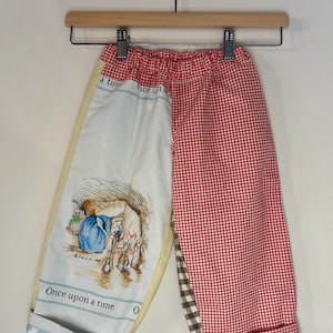 May include: Children's pants with a patchwork design. One leg features a red and white gingham pattern, while the other has a cream-colored panel with a storybook illustration and the words "Once upon a time." The cuffs are blue with white stars.