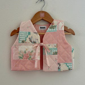 May include: A quilted vest with a patchwork design. The vest features a pink and white gingham pattern, floral and striped fabric patches, and a pink bow closure. The vest is hanging on a wooden hanger with the text "Skylark".