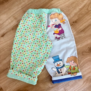 May include: Handmade children's trousers with a whimsical design. One leg has a light green floral pattern, the other has illustrations of a girl with soap, a snowman, and another girl. The trousers have a mint green waistband and cuffs.