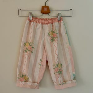 Könnte beinhalten: Babyhose mit rosa und weißen Streifen und floralen und Band-Akzenten. Die Hose hat einen rot-weiß karierten Bund und ist an den Knöcheln umgeschlagen. Sie hängt an einem Holzkleiderbügel.