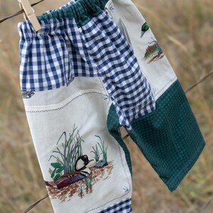 May include: Pants featuring a patchwork design with cream fabric panels displaying duck and marsh illustrations, alternating with navy blue and white gingham check sections, and a solid dark green textured fabric panel.