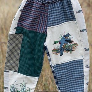 May include: Patchwork pants featuring a variety of blue, green, and plaid fabric panels. The design includes printed illustrations of ducks in flight and nesting in reeds. The waistband is elasticized and features a white label reading Skylark.