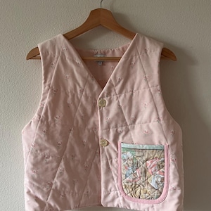 Puede incluir: Un chaleco acolchado rosa con estampado floral y un bolsillo de parche con una ilustración caprichosa. El chaleco tiene cierre de botón y un patrón de acolchado en forma de diamante.