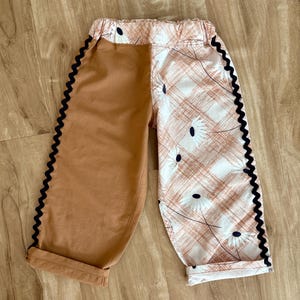 May include: Two-toned pants with a brown solid color on one side and a white and peach plaid pattern with floral accents on the other. Black zigzag trim lines the outer seams. The pants have a rolled hem and an elastic waistband.