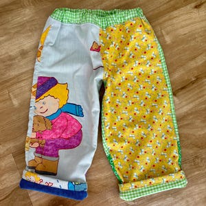 May include: Children's patchwork trousers with a cartoon illustration on one leg and a dog pattern on the other. The design includes green gingham trim and blue cuffs. The trousers are a vibrant mix of colours.