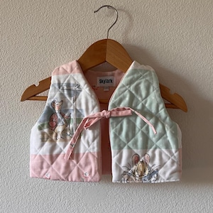 Puede incluir: Un chaleco acolchado rosa y verde con un estampado de Peter Rabbit en un lado y un ratón leyendo un libro en el otro. El chaleco tiene un cierre de corbata y una etiqueta "Skylark".