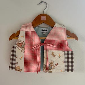 Puede incluir: Un chaleco acolchado con temática de Peter Rabbit, con cuadros rojos, cuadros marrones y tela color crema con ilustraciones. El chaleco tiene un cierre de lazo y está colgado de una percha de madera.