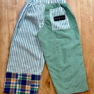 Puede incluir: Un pantalón con diseño de patchwork que presenta un patrón de rayas verticales azules y blancas en un lado y un patrón de microcuadros verdes y blancos en el otro. El dobladillo tiene un panel de cuadros multicolor y un bolsillo muestra una etiqueta Ralph Lauren.