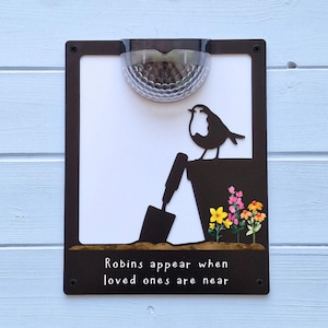 May include: A black metal garden sign with a solar light. The sign features a bird silhouette, a trowel, and colourful flowers. The text "Robins appear when loved ones are near" is displayed at the bottom of the sign.