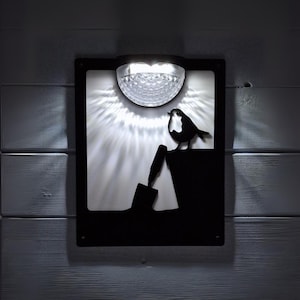 May include: Black metal wall art depicting a bird silhouette above a shovel. A solar-powered light is positioned above, casting a bright, starburst pattern. The art is mounted on a white wooden wall, creating a striking contrast.
