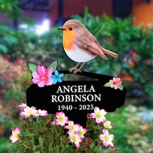 May include: Black metal garden memorial with a robin bird perched on top. The memorial has a white inscription that reads "ANGELA ROBINSON 1940 - 2025" and is decorated with pink and blue flowers.